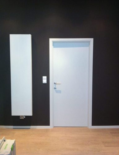 Une porte blanche sur un mur noir, avec un radiateur vertical à gauche.